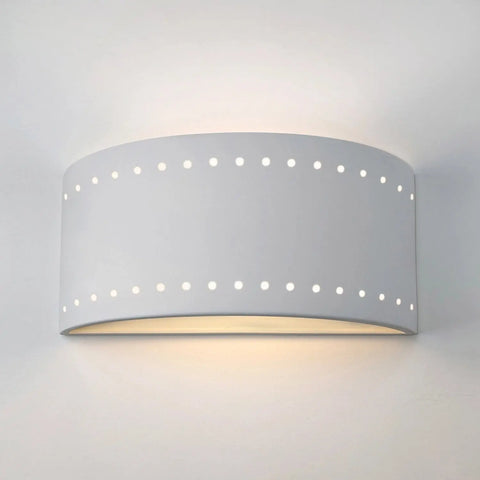 The Syros Sconce by A19 Artisan Lighting is a modern, handcrafted white ceramic wall light made in California, featuring a curved design with two rows of circular perforations that softly emit light from the top and bottom.