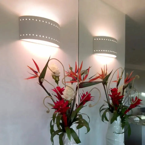 A vase with red and white tropical flowers sits in front of a mirror. Above, the Syros Sconce by A19 Artisan Lighting, handcrafted in California, softly illuminates the white wall with its delicate perforated ceramic design.