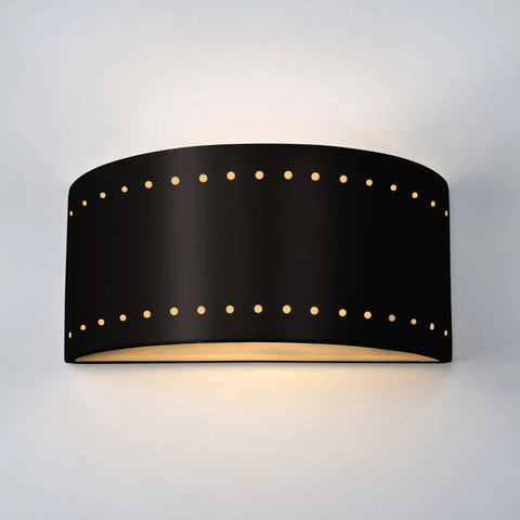 The Syros Sconce by A19 Artisan Lighting is a modern black ceramic wall light, handcrafted in California, featuring two rows of small holes and emitting soft up-and-down light on a white wall.