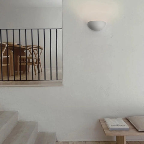 A minimalist interior with light walls and stairs, a black metal railing, wooden dining furniture, a handcrafted USA bench with stacked magazines, and the Thera Sconce by A19 Artisan Lighting—modern wall lighting handmade in California.