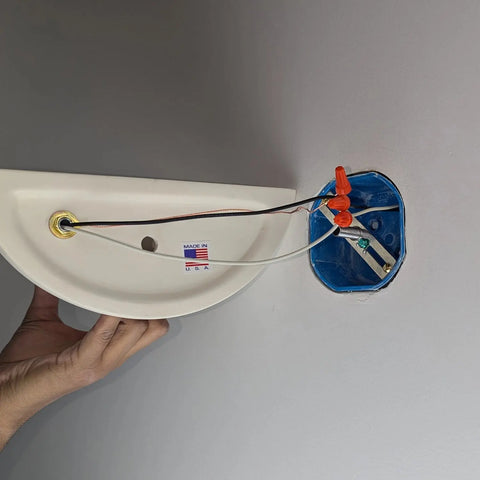 A person prepares to install the Thera Sconce, a customizable wall light by A19 Artisan Lighting, handcrafted in California, holding it with exposed wiring next to a blue electrical box mounted in the wall.