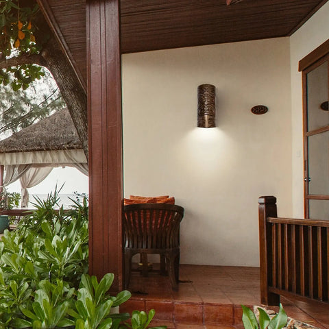 A shaded porch features a wooden chair and table, tropical plants in front, and Modern USA-Made Wall Lighting – Tiki Downlight Sconce by A19 Artisan Lighting, with a thatched roof gazebo partially visible in the background.