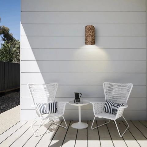 Two white wicker chairs with striped cushions face a small round table with a black pitcher, set on a wooden deck against a white paneled wall, accented by the Tiki Downlight Sconce by A19 Artisan Lighting, handcrafted in California.