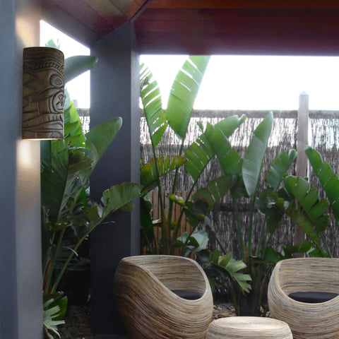Modern patio with tropical plants, two sculpted wood chairs, a side table, and a fence; the left wall features the Modern USA-Made Wall Lighting – Tiki Sconce by A19 Artisan Lighting, handcrafted in California with energy-efficient LED smart bulbs.