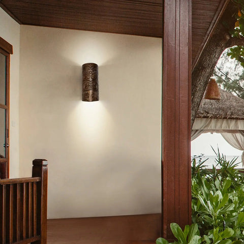 The Modern USA-Made Wall Lighting – Tiki Sconce by A19 Artisan Lighting, handcrafted in California and fitted with energy-efficient LED smart bulbs, illuminates a cream porch wall near wooden pillars and lush greenery.