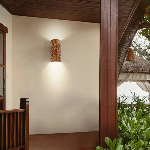 The Modern USA-Made Wall Lighting – Tiki Sconce by A19 Artisan Lighting, handcrafted in California, is installed on a beige exterior wall near a wooden porch, with plants and a thatched-roof structure visible in the background.