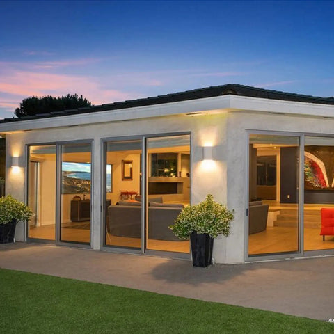 Contemporary single-story home at dusk with potted plants and spacious glass doors, featuring illuminated Modern USA-Made Wall Lighting – Tilos Sconce by A19 Artisan Lighting, handcrafted in California—stylish interiors glow with sunset skies.