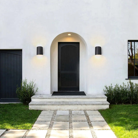 A modern house entrance with a black door under an arched porch is flanked by two Modern USA-Made Tilos Sconces, handcrafted in California by A19 Artisan Lighting, plus stone steps, green lawn, shrubs, and a paved walkway leading to the door.