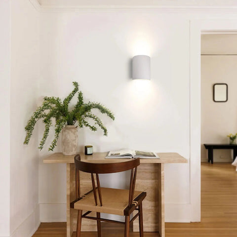 A wooden chair sits at a small desk with an open book, greenery, and a candle. Above, the Tilos Sconce by A19 Artisan Lighting—modern and handcrafted in California—casts a warm glow. A doorway leads to another room with a bench and wall art.