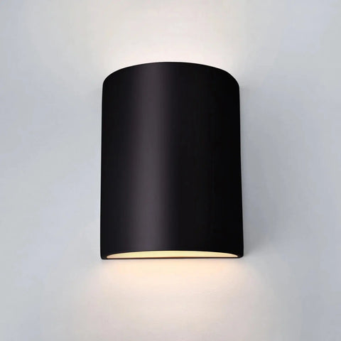 The Modern USA-Made Tilos Sconce by A19 Artisan Lighting, handcrafted in California, features a sleek black cylindrical design that glows softly from top and bottom when mounted on a plain white wall.