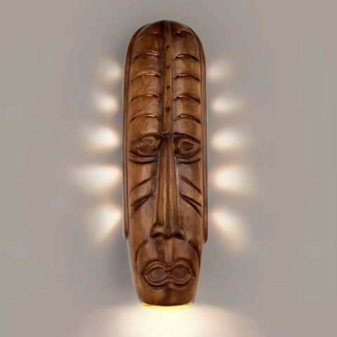 A tribal mask-shaped wall sconce with an abstract design, emitting light from its open bottom.