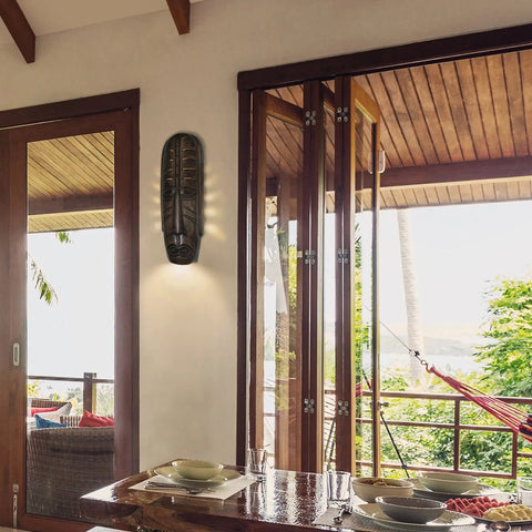 A tropical dining area with a wooden table and glassware overlooks a porch with a hammock. LED smart bulbs create cozy lighting, while the A19 Artisan Lighting Tribal Mask Sconce hangs near sliding doors framing lush greenery.