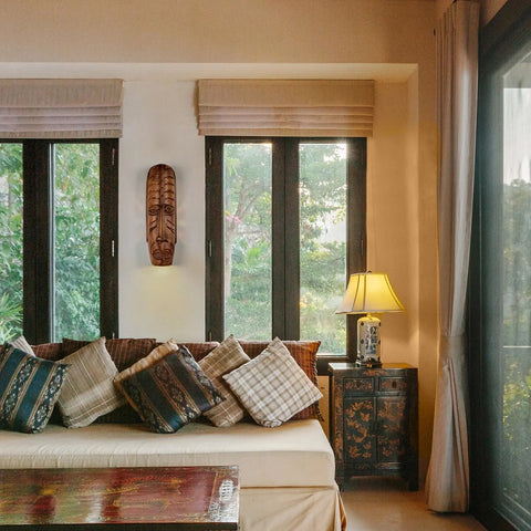 A cozy living room features a cushioned sofa with patterned pillows, a Modern USA-Made Wall Lighting – Tribal Mask Sconce by A19 Artisan Lighting, large windows with greenery, LED smart bulbs in a lamp, and an ornate cabinet beside the sofa.