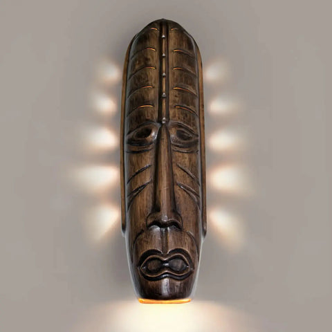 A tribal mask-shaped wall sconce with an abstract design, emitting light from its open bottom.