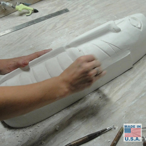 An artisan works on a Modern USA-Made Wall Lighting – Tribal Mask Sconce by A19 Artisan Lighting, handcrafted in California and surrounded by sculpting tools. The Made in USA label is visible on the worktable.