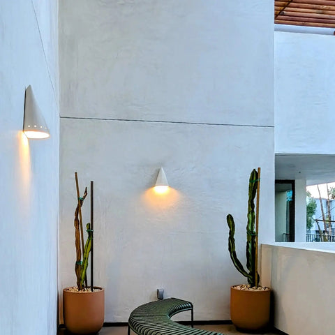 A modern balcony with white walls, two tall potted cacti, a curved green bench, and ceramic lighting featuring the Modern USA-Made Zealandia Downlight Sconce by A19 Artisan Lighting. Natural light enters from the right with part of a wooden ceiling above.