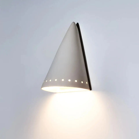 The Modern USA-Made Zealandia Downlight Sconce by A19 Artisan Lighting is handcrafted in California, featuring a cone-shaped ceramic design with circular perforations that cast a warm glow onto any white wall.