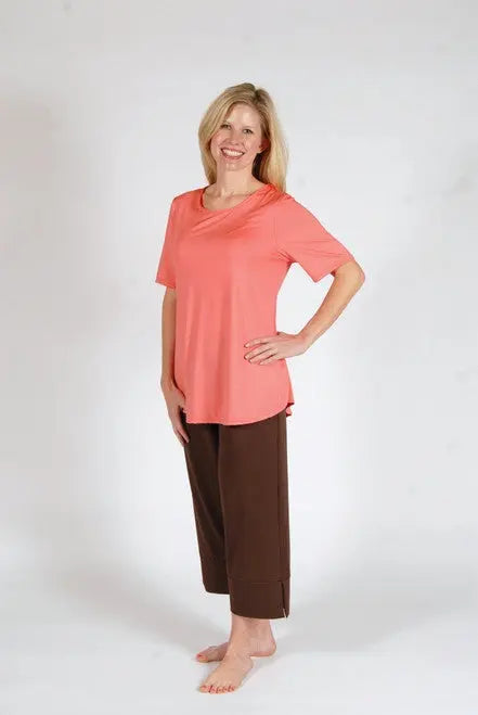 A smiling blonde woman stands barefoot on a white background, wearing Goodnighties Cropped Pant in brown and a loose coral short-sleeve top, both crafted from anti-microbial fabric.