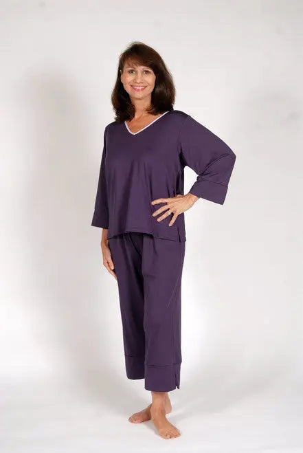 A smiling woman with shoulder-length brown hair stands barefoot on a white background, wearing the Goodnighties Cropped Pant and a loose-fitting, long-sleeve purple top made from moisture-wicking sleepwear fabric.