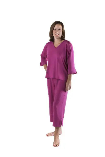 A woman with shoulder-length brown hair stands barefoot, smiling in Goodnighties magenta long-sleeved top and matching Cropped Pant sleepwear, known for moisture-wicking comfort, against a white background.