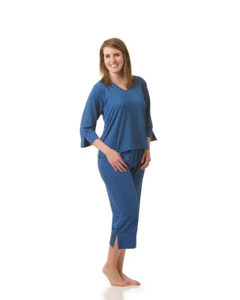 A woman stands barefoot, smiling, in Goodnighties Cropped Pant—a loose-fitting blue pajama set with three-quarter sleeves and cropped sleep pants made from anti-microbial fabric—against a plain white background.