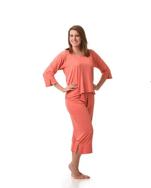 A woman stands barefoot on a white background, smiling with her hands on her hips. She is wearing Goodnighties Cropped Pant, a loose-fitting, coral-colored pajama set featuring moisture-wicking sleepwear for comfort.