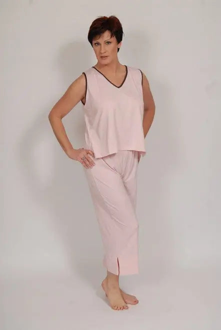 A woman with short dark hair stands barefoot against a plain background, wearing Goodnighties Cropped Pant—light pink sleeveless pajamas made from anti-microbial fabric—with one hand on her hip and a neutral expression.