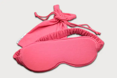 The Goodnighties Sleep Mask With Pouch features a pink handcrafted mask with an elastic strap, displayed in front of a matching drawstring pouch on a light gray background.