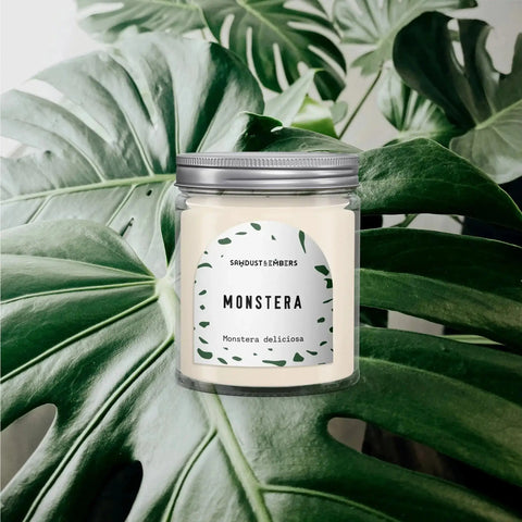 The Monstera - Signature Candle by Sawdust & Embers rests on green monstera leaves, featuring a label with leaf illustrations and “Monstera deliciosa” beneath the name—a stylish home fragrance accent.