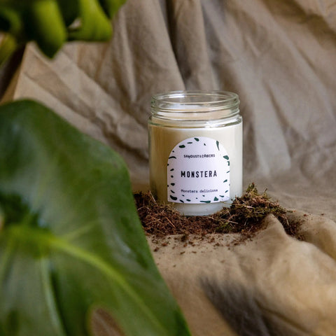 The Sawdust & Embers Monstera - Signature Candle, crafted from soy wax in a glass jar, rests on beige fabric with scattered soil beside a lush monstera leaf, adding elegant home fragrance amidst soft draped backgrounds.
