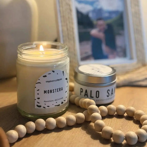 A glowing Sawdust & Embers Monstera - Signature Candle fills the air with fragrance, sitting beside a Palo Santo tin and wooden bead garland on a wooden surface, with a blurred framed photo in the background.