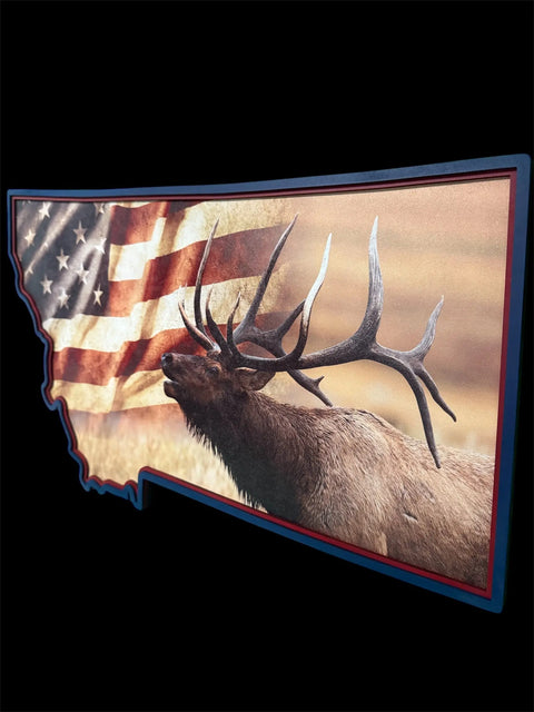 The Montana Elk Flag Sign by B. James Design & Woodwork features a bull elk silhouette inside the Montana shape, set against a waving American flag and blurred natural background—a unique tribute to Montana wildlife.