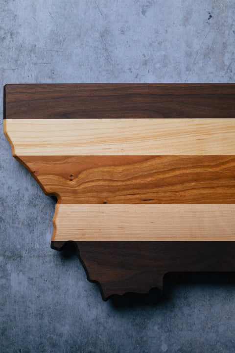 The Montana by Montana Block is a handcrafted wood cutting board shaped like Montana with three striped wood tones, displayed on a gray textured surface—a personalized kitchen gift for any occasion.