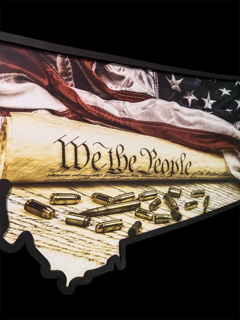 A curled U.S. Constitution with "We the People" visible lies beside scattered bullets and a partially visible American flag, highlighting the Montana We the People Sign by B. James Design & Woodwork against a black backdrop.