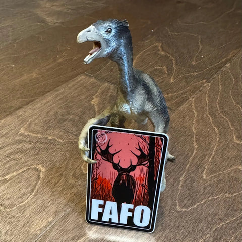 A dinosaur figurine stands on a wooden floor, holding a rectangular sign featuring the American Lictor Sticker: Moose - FAFO, which displays a bold, stylized moose head and the letters FAFO underneath.