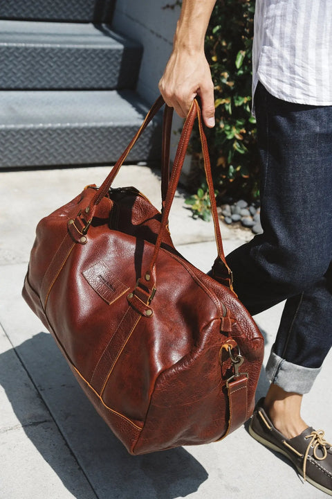 A person in cuffed jeans, a striped shirt, and boat shoes walks outside, carrying the Rogue Industries Mount Blue Leather Duffle by its handles.