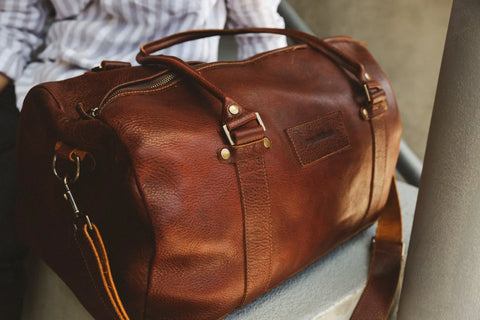 A close-up of the Mount Blue Leather Duffle by Rogue Industries, crafted from American bison leather with brass hardware and a shoulder strap, sits on concrete. A person in a striped shirt appears partially in the background.