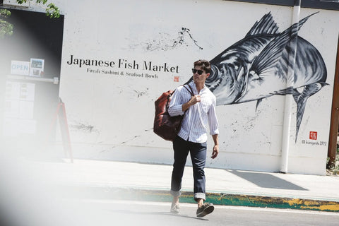 A man wearing sunglasses, a striped shirt, and rolled-up pants carries the Mount Blue Leather Duffle by Rogue Industries past a wall mural of a large fish and a sign that reads Japanese Fish Market: Fresh Sashimi & Seafood Bowls.