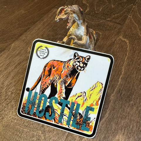 A toy dinosaur stands on wood behind the American Lictor Sticker: Mountain Lion - Hostile, which shows a bold cougar and HOSTILE text like a stealth patch, surrounded by rocks and grass.