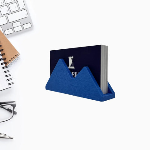 The Chatelet Manufacturing Mountain Silhouette Design Business Card Holder stylishly displays business cards on a white desk, serving as a unique desk accessory alongside a keyboard, spiral notebook, pen, and eyeglasses.
