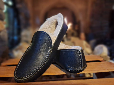 A pair of black BBH Nash Island Slipper - Lone Sole sheepskin-lined leather slippers sits on a wooden surface, with a warm, blurred fireplace in the background.