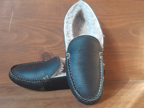 A pair of BBH Nash Island Slipper - Lone Sole, black leather with visible stitching and soft sheepskin lining, are showcased on a wooden surface—one standing upright, the other lying flat.