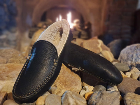 A pair of BBH Nash Island Slipper - Lone Sole, black sheepskin-lined leather slippers with handsewn details, rests on stones before a glowing fireplace.