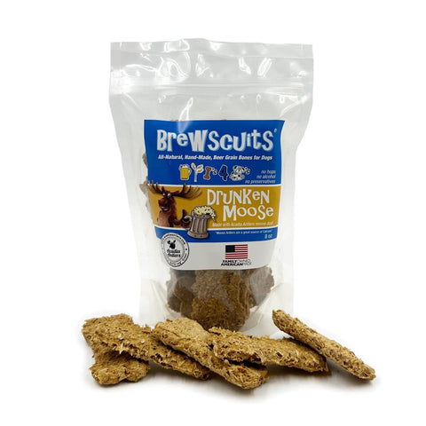 A Brewscuits Drunken Moose dog treats bag stands upright, featuring a moose and beer mug image, with brown textured treats scattered in front; packaging highlights the addition of moose antler dust.