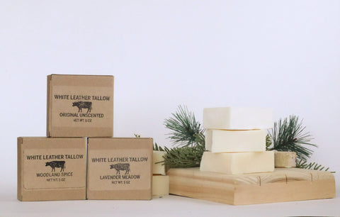 Three labeled White Leather Tallow Handmade Tallow Soap boxes are stacked beside a wooden tray with four plain bars and pine sprigs, all set against a white background. Made with premium grass-fed beef tallow.