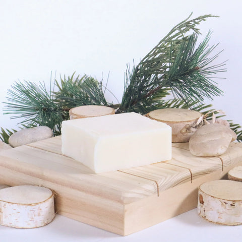 A bar of White Leather Tallow’s Handmade Tallow Soap rests on a wooden platform, surrounded by birch slices, stones, and pine branches against a white background.