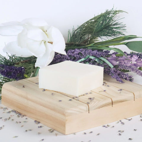 A white bar of Handmade Tallow Soap by White Leather Tallow sits on a wooden dish, with lavender buds, sprigs of lavender and pine, and a white flower in the background.