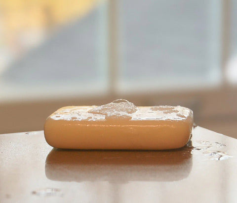 A wet bar of White Leather Tallow’s Handmade Tallow Soap, covered in bubbles, rests on a smooth surface with water droplets around it. The blurred background features soft light and gentle, indistinct shapes.
