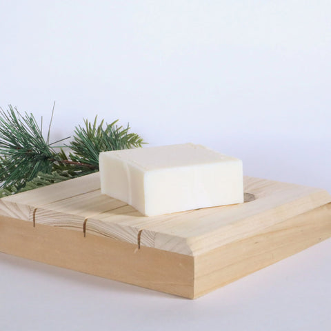 A bar of White Leather Tallow’s Handmade Tallow Soap sits on a light wooden dish with a pine branch behind it, set against a plain white background.