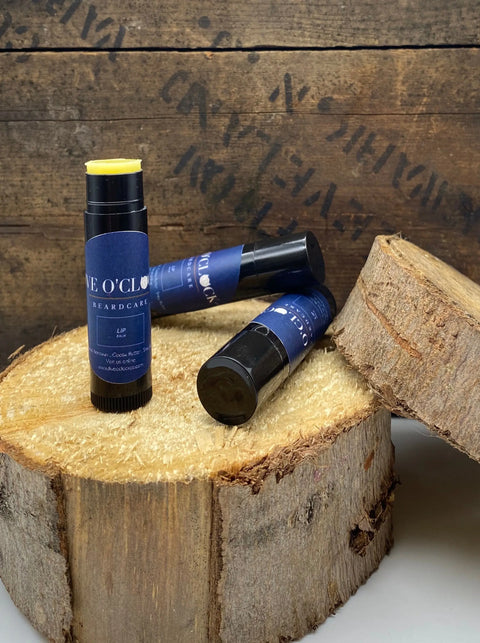 Three dark blue Lip Balm tubes from Five O Clock Co. rest on rustic wooden slices, one uncapped to show the yellow balm. Ideal for hydration, theyre set against a textured, unfinished wood background.